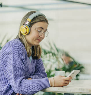 Lächelndes Mädchen, das über drahtlose Kopfhörer in der Schulcafeteria sitzt und Musik hört - VSNF00755