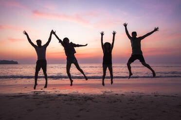 Freunde springen mit erhobenen Armen am Strand bei Sonnenuntergang - IKF00289