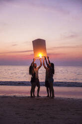 Friends burning paper lantern standing at beach on sunset - IKF00294