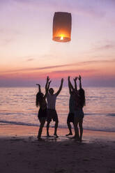 Friends releasing lit paper lantern standing at beach on sunset - IKF00295