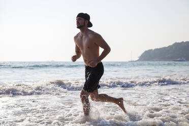 Shirtless man running in water at beach - IKF00336