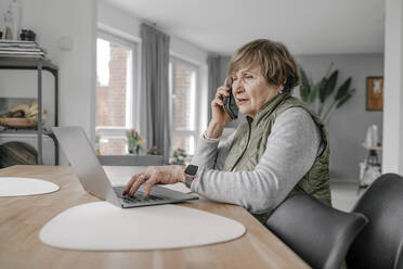 Ältere Frau, die zu Hause einen Laptop benutzt und mit ihrem Smartphone telefoniert - OGF01295