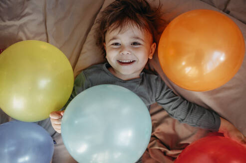 Glücklicher Junge auf dem Bett liegend inmitten von Luftballons - ANAF01272