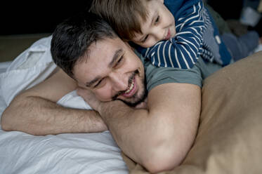 Happy man lying with son on bed at home - ANAF01277