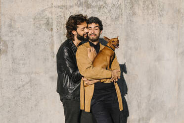 Schwuler Mann umarmt seinen Freund mit Hund vor einer Betonwand - MMPF00742