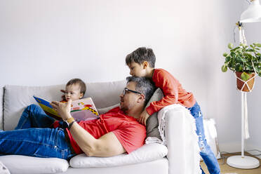 Father reading book with sons on sofa in living room at home - JJF00891