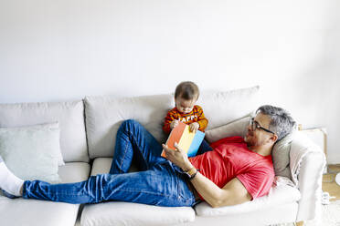 Father lying on sofa with son and book at home - JJF00893