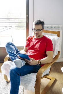 Mann mit Tablet-PC auf einem Stuhl sitzend zu Hause - JJF00896