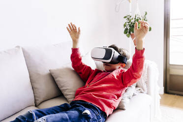 Junge mit erhobenen Armen, der ein Virtual-Reality-Headset trägt und zu Hause auf dem Sofa liegt - JJF00897