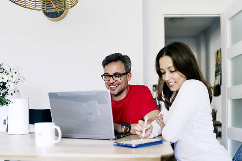 Mann arbeitet an einem Laptop und sitzt neben einer Frau, die zu Hause einen Tablet-PC benutzt - JJF00901