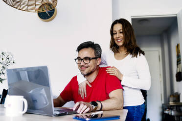 Frau mit Arm um Mann, der zu Hause am Laptop arbeitet - JJF00904