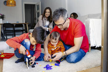 Father sitting by sons playing with toys in living room at home - JJF00932