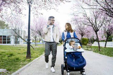 Vater und Mutter gehen mit Kinderwagen auf dem Fußweg im Park spazieren - JJF00950