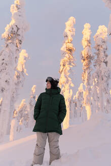 Junge Frau in grüner Jacke vor schneebedeckten Bäumen stehend - LHPF01525