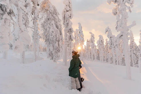 Junge Frau mit Snowboard auf einem schneebedeckten Feld bei Sonnenaufgang - LHPF01529