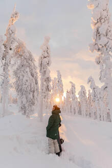 Junge Frau mit Snowboard auf einem schneebedeckten Feld bei Sonnenaufgang - LHPF01530