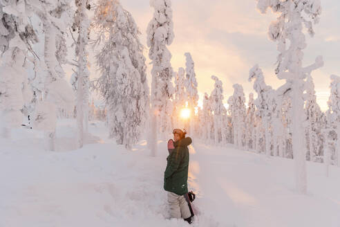 Frau mit Snowboard auf einem schneebedeckten Feld bei Sonnenaufgang - LHPF01531