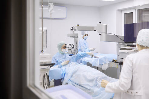 Augenarzt bei einer Augenoperation im Operationssaal, gesehen durch ein Glasfenster - SANF00091