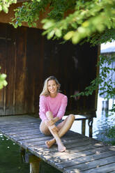 Smiling woman sitting on boardwalk at lake - PNEF02762