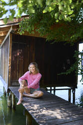 Smiling woman sitting outside house on boardwalk at lake - PNEF02769