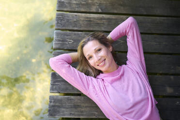 Smiling woman with hands behind head lying on boardwalk - PNEF02775