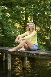 Smiling woman sitting on boardwalk at lake - PNEF02785