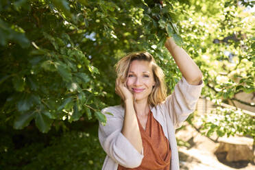 Lächelnde Frau mit Hand im Haar unter einem Baum stehend - PNEF02790