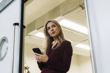 Nachdenkliche Geschäftsfrau hält Mobiltelefon in der Nähe der Tür in einer Fabrik - JOSEF18694