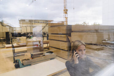 Businesswoman talking on mobile phone in carpentry factory seen through glass - JOSEF18752