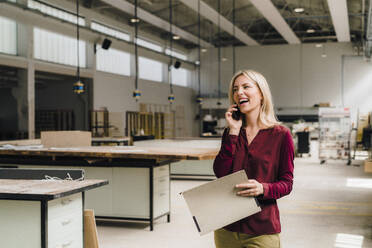 Happy businesswoman talking on smart phone holding file in factory - JOSEF18824