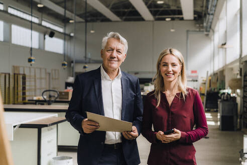 Smiling colleague with senior businessman holding document in factory - JOSEF18833