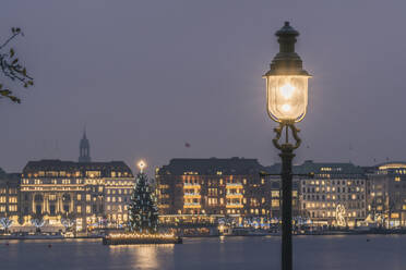 Germany, Hamburg, Street light glowing in front of Alster Lake at dusk with Christmas tree in background - KEBF02707