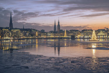 Deutschland, Hamburg, Eisschwimmen auf der Alster in der Abenddämmerung mit der Skyline der Stadt und leuchtenden Weihnachtsbäumen im Hintergrund - KEBF02715