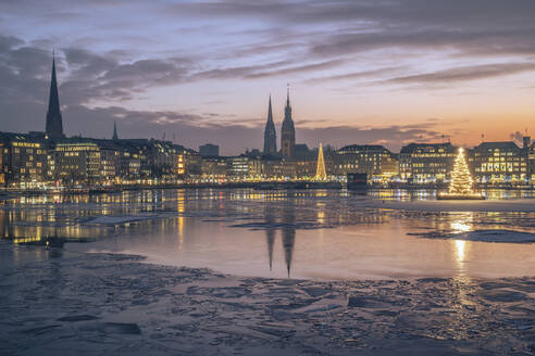 Deutschland, Hamburg, Eisschwimmen auf der Alster in der Abenddämmerung mit der Skyline der Stadt und leuchtenden Weihnachtsbäumen im Hintergrund - KEBF02715