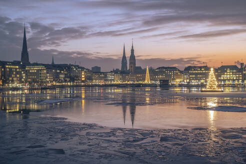 Germany, Hamburg, Ice floating in Alster Lake at dusk with city skyline and glowing Christmas trees in background - KEBF02715