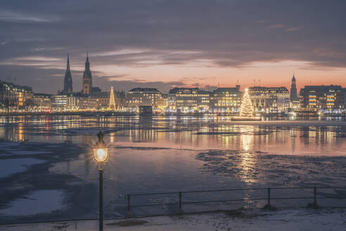 Germany, Hamburg, Ice floating in Alster Lake at dusk with city skyline and glowing Christmas trees in background - KEBF02716
