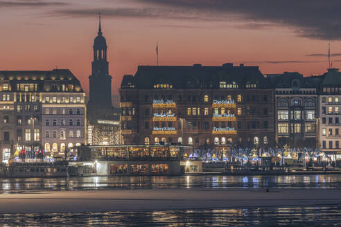 Germany, Hamburg, Christmas decorations hanging on city buildings at dusk with Alster Lake in foreground - KEBF02717