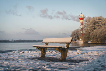 Deutschland, Hamburg, Leere Bank am schneebedeckten Elbufer mit Leuchtturm Wittenbergen im Hintergrund - KEBF02744