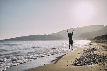 Cheerful woman walking with arms raised near shore at beach - ANNF00142