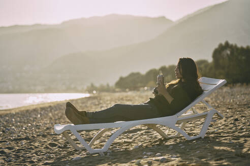 Frau auf Liegestuhl liegend mit Thermoskanne am Strand - ANNF00144