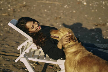 Lächelnde Frau auf Liegestuhl liegend und Hund streichelnd am Strand - ANNF00145
