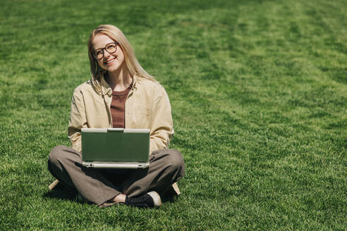 Glücklicher Freiberufler mit Laptop im Schneidersitz auf Gras im Garten - VSNF00764