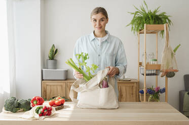 Smiling woman removing celery from reusable bag at home - ALKF00253