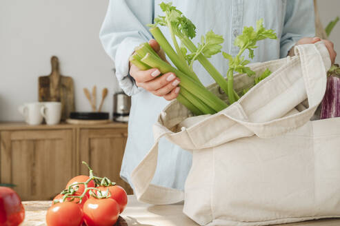 Woman holding fresh green celery in reusable bag at home - ALKF00254