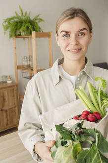 Lächelnde Frau mit einer Tüte mit Bio-Gemüse und Obst zu Hause - ALKF00272