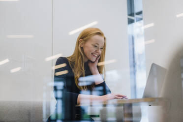 Lächelnde Geschäftsfrau, die an einem Laptop arbeitet und in einer Bürozelle sitzt - JOSEF18938