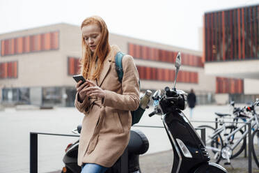 Junge Frau, die ein Smartphone benutzt und sich auf einen Motorroller stützt - JOSEF18977