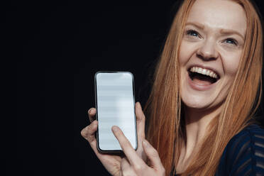 Junge Frau lachend und hält Smartphone vor schwarzem Hintergrund - JOSEF19020