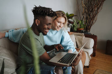 Smiling man pointing at laptop by girlfriend sitting on sofa at home - MDOF01064