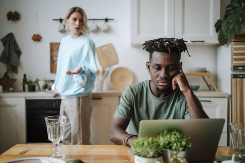 Stressed man watching laptop with woman arguing in background at home - MDOF01099